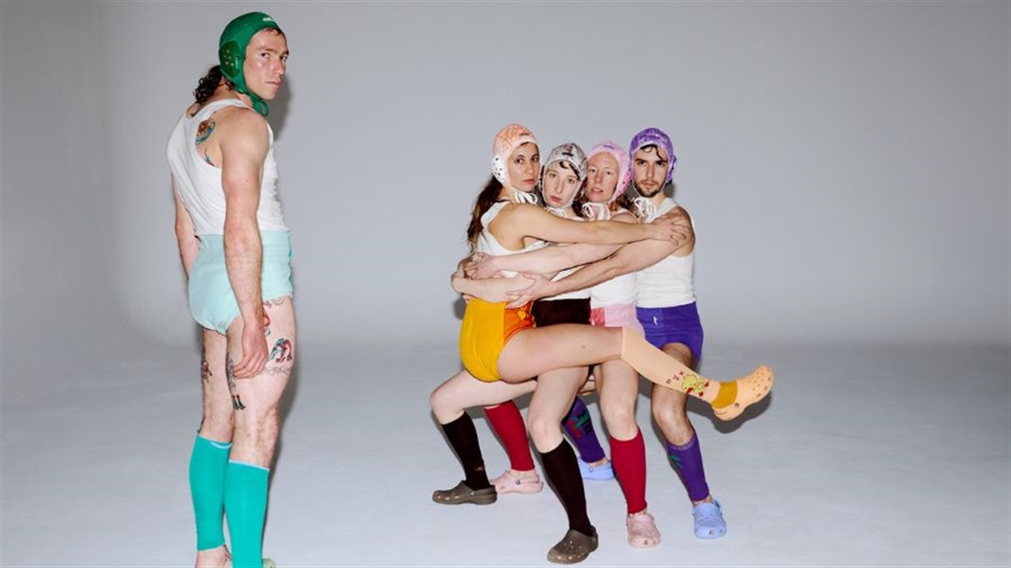 Four people wearing colourful wrestling outfits squat one in front of the other. The person on the left of the cluster sits on legs facing in the other direction. A person also wearing a colourful wrestling outfit stands to the left of the group.