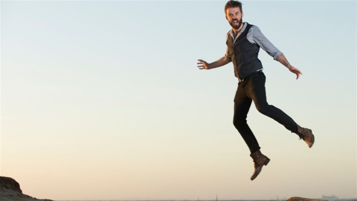 A man with short dark spikey hair wears dark trousers, a dark vest and a light buttoned shirt with the sleeves rolled up. He jumps in the air amongst a seaside landscape.