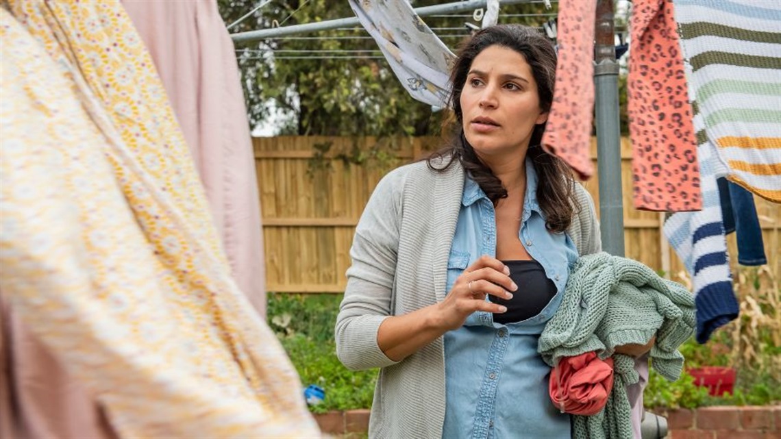 A woman with long dark hair stands under a clothes line where linens and clothes are drying. She stares intently towards her right. There is a beige yard fence behind her.