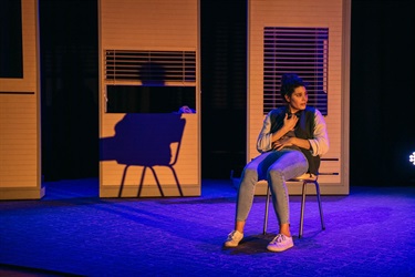 A woman with dark curly hair tied up into a high bun sits on a chair. She wears blue jeans and a letter jacket. She holds her chest as if concerned and looks over her left shoulder.