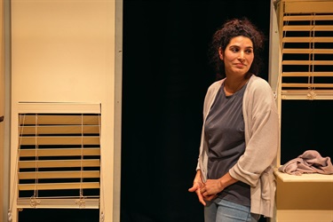 A woman with dark curly hair pulled back into a high bun leans on a window frame. She wears a grey tshirt, a beige cardigan and blue jeans. Her hands are folded in front of her waist.