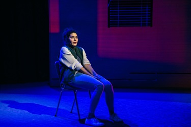 A woman with dark curly hair sits on a chair off centre of a stage. She wears a letter jacket and blue jeans. Her hands are tucked between her legs as she stares with concern into the horizon.