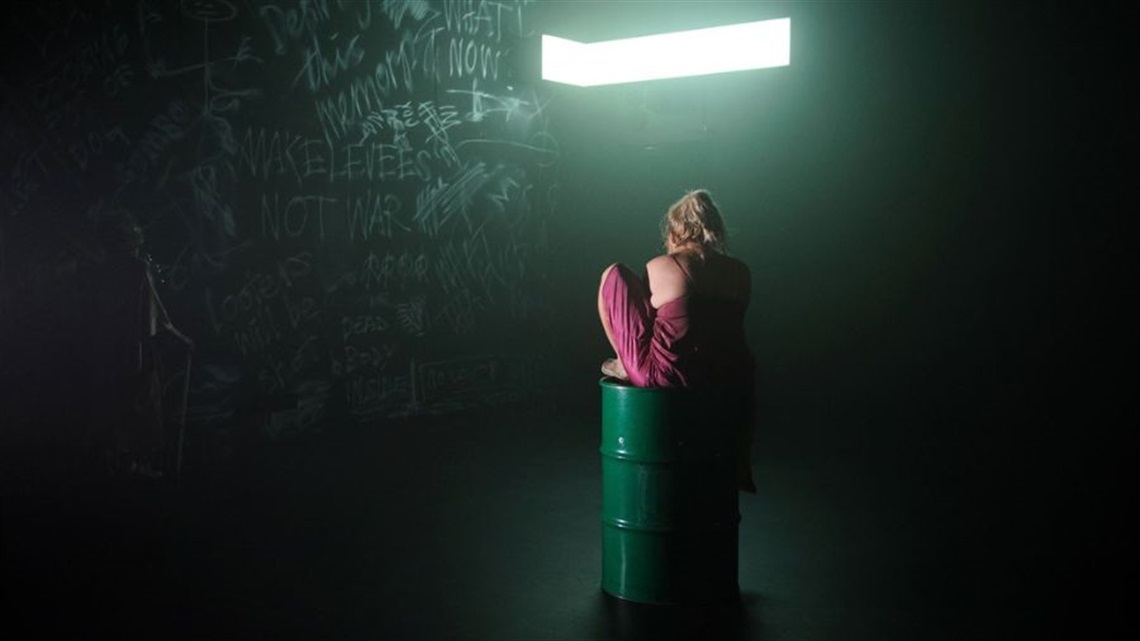 A person sits on top of an oil barrel inside a dark room with white chalk writing on a black wall.