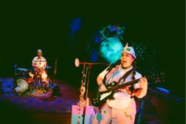 A woman wearing a funky hat stands centre stage behind a microphone. She is playing the guitar and singing. A person sits behind her on the left behind a drum kit.
