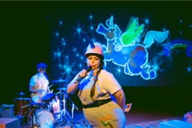 A woman wearing overalls and a unicorn hat stands in profile with a microphone to her mouth. Her lips pout. A large unicorn image is behind her.