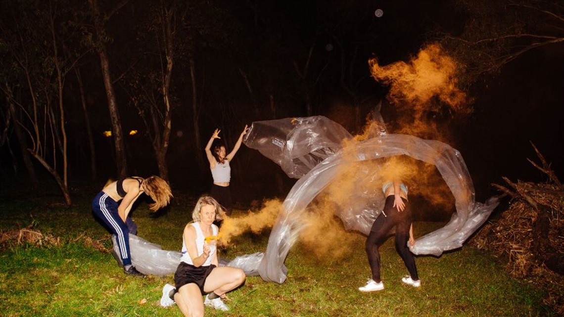 Four female dancers jump, kick and crouch around a wooded area at night. Smoke billows between them. A long train of plastic is held by two people.