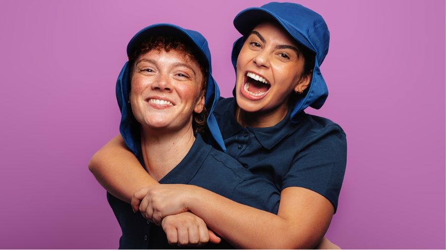 A person hugs another from behind. They both wear dark-blue collared t-shirts and blue capped hats.