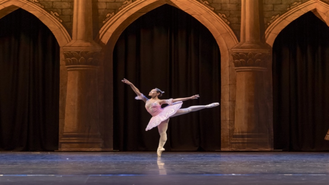 A ballerina wearing a pink tutu stands on one leg, the other leg stretched outwards behind her and her right arm stretched in the air. Behind her is a painted backdrop of stone-cut arches.