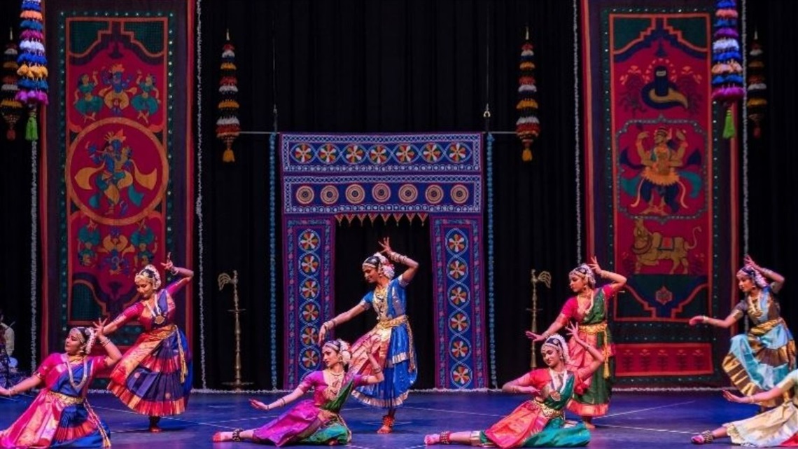 A group of traditional Indian dances spread across a proscenium stage. Half stand, and half kneel on the ground.