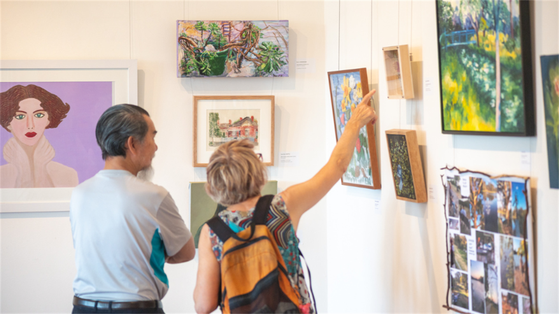 Two people view a wall of salon hung style artworks