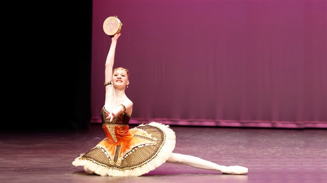 A female ballerina wearing a Renaissance like tutu and headpiece, lounges on her right side on a stage. Her right arm is raised over her head and her feet are pointed to her right. Behind her is a mauve curtain. 