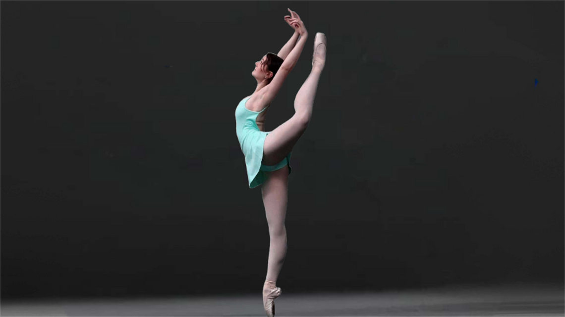 A ballerina wearing a light blue leotard and pointed ballerina shoes stands on one leg with arms raised in the air over her head.