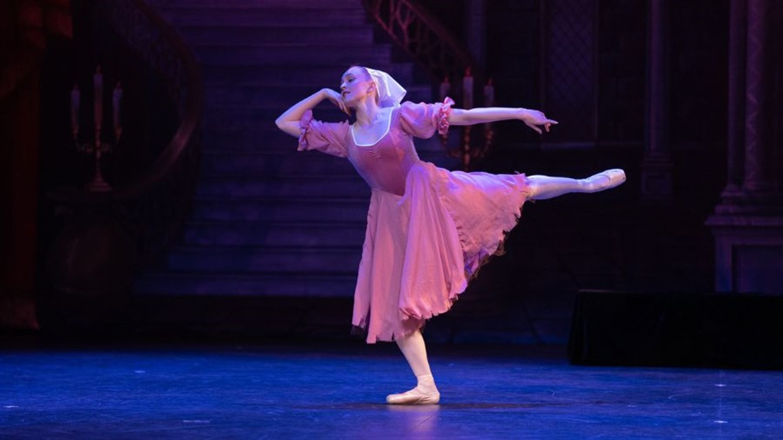 A ballerina wearing a light pink long dress and a beige headscarf on her head stands on one foot with the other foot extended behind.