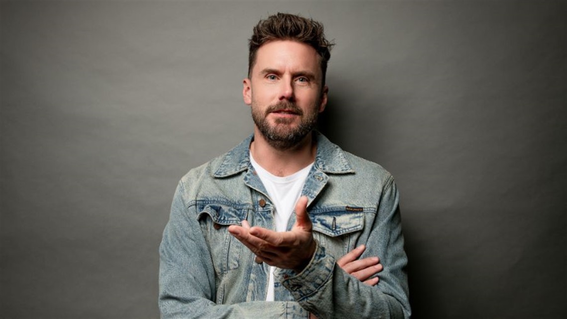 A man with short dark hair and a beard wears a white tshirt under a denim jacket. He folds his right arm across his chest and holds his left hand out.