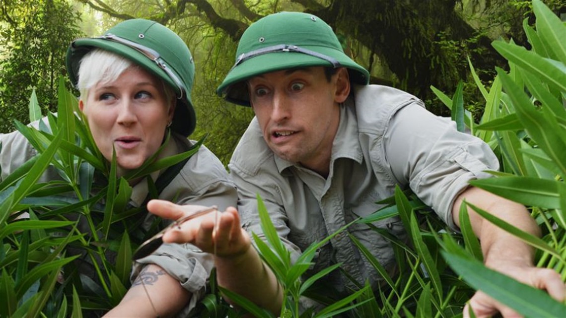 Two people dressed in safari clothes and green hats peer out from behind tall green grass.