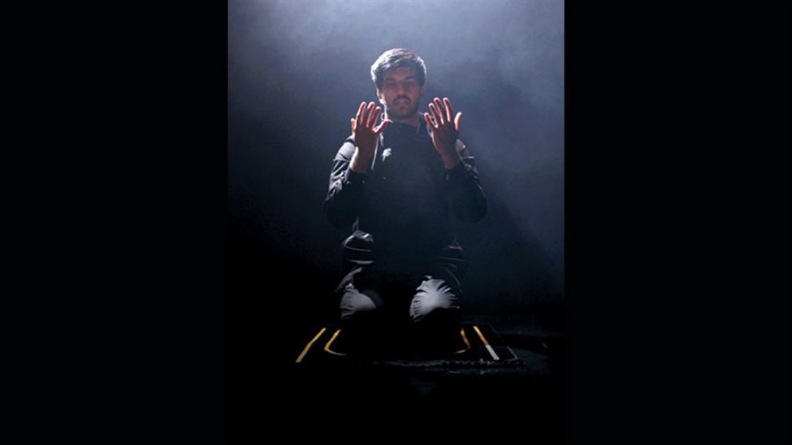 A man with short dark hair and a short beard wears an all black outfit and sits on his legs on a rug. He holds out his hands in front looking at his palms.