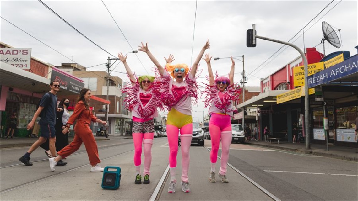 Three people wearing white and pink outfits and coloured hair jump with their arms raised in the middle of a public street. To their left a group of people walk by.