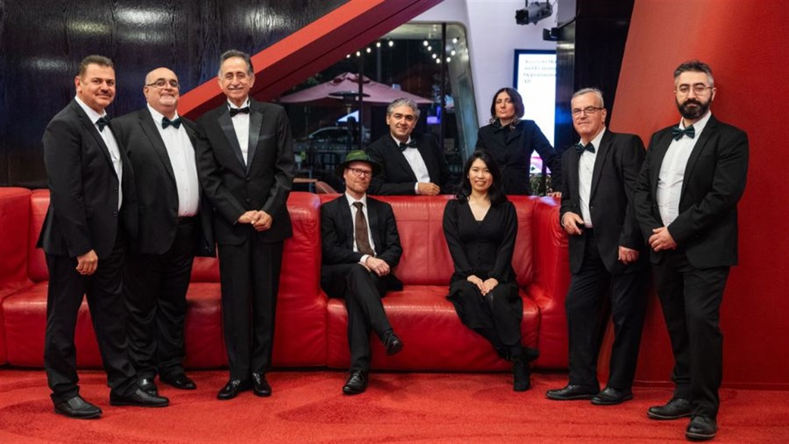 A group of men and women wearing black suits with white buttoned down shirts and black cocktail dresses gather around a red leather sofa. 