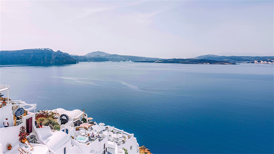 A view of white stone houses along a cliffside facing a deep blue ocean.