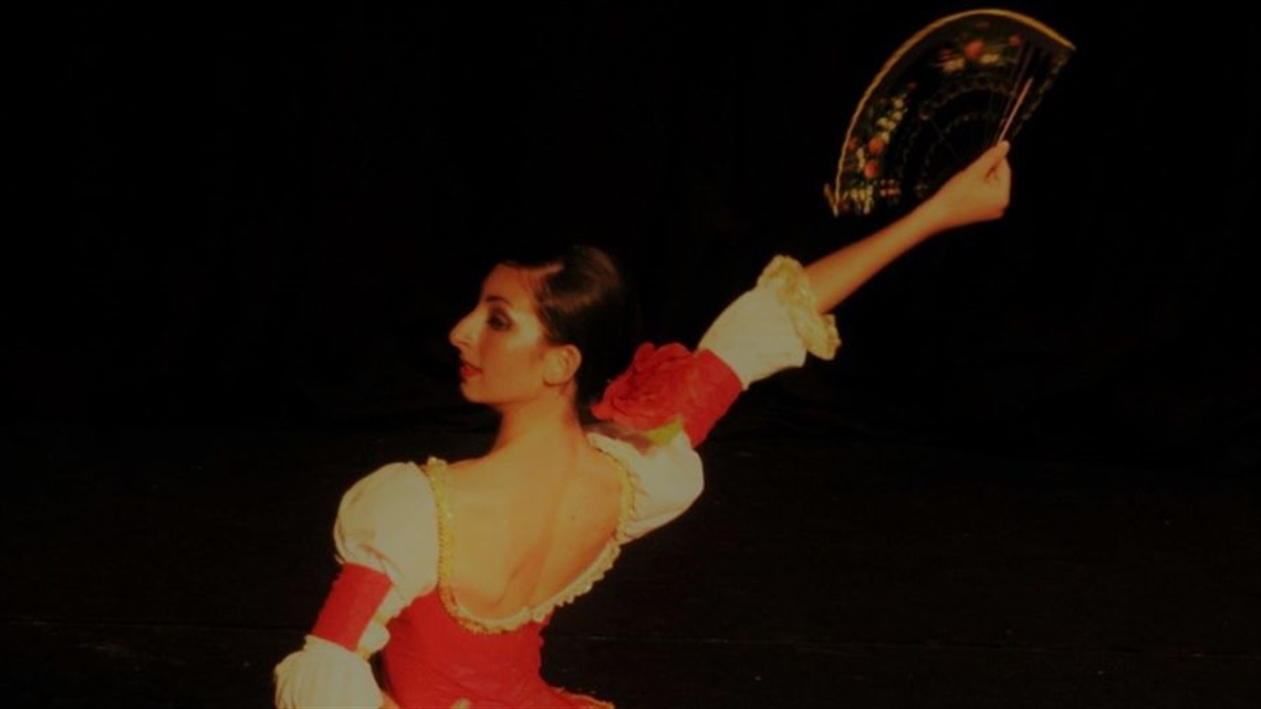 A woman with dark hair pulled back into a bun wears a red top that falls over her shoulders. Her back is facing us with her head turned in profile, and she raises her right arm in the air while holding a black fan.