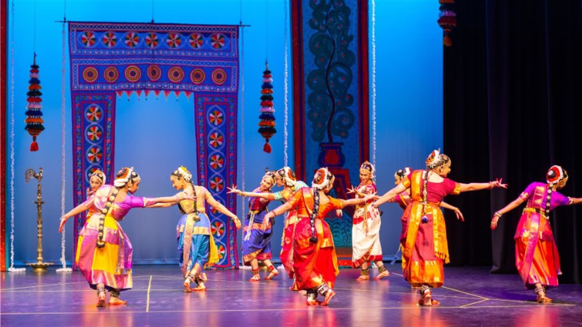 A group of female Indian dancers are scattered across a proscenium stage. They wear traditional Indian garments including headdresses, in various colours. Behind them is a backdrop of a temple entrance. 