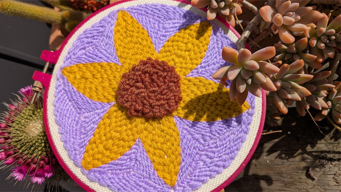 A colouful punch needle project of a flower in yellow, purple and brown