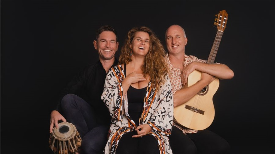 A woman and two men sit side by side one another. The man on the right holds a guitar, while the man on the left holds a bongo drum.