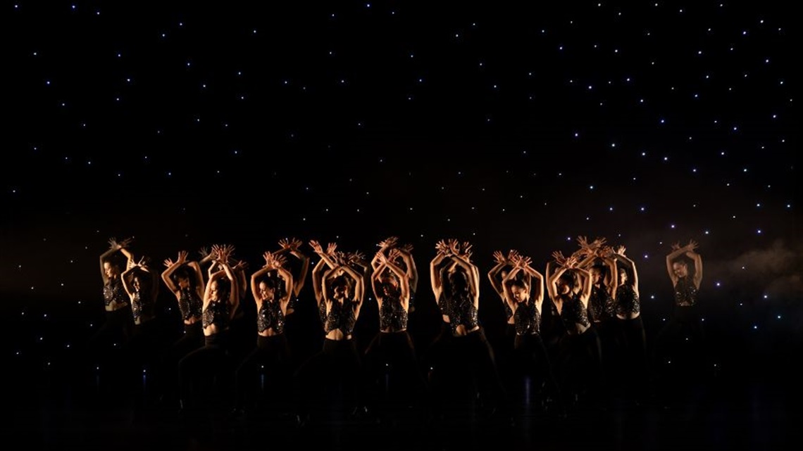 A group of female dancers wearing dark pants and dark sparkly tops hold their hands in the air in front of a tinkling backdrop.