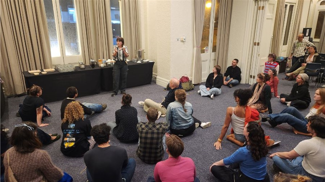 A person with short dark hair wears dark trousers and a striped shirt stands in front of a crowd of people sitting on the floor.