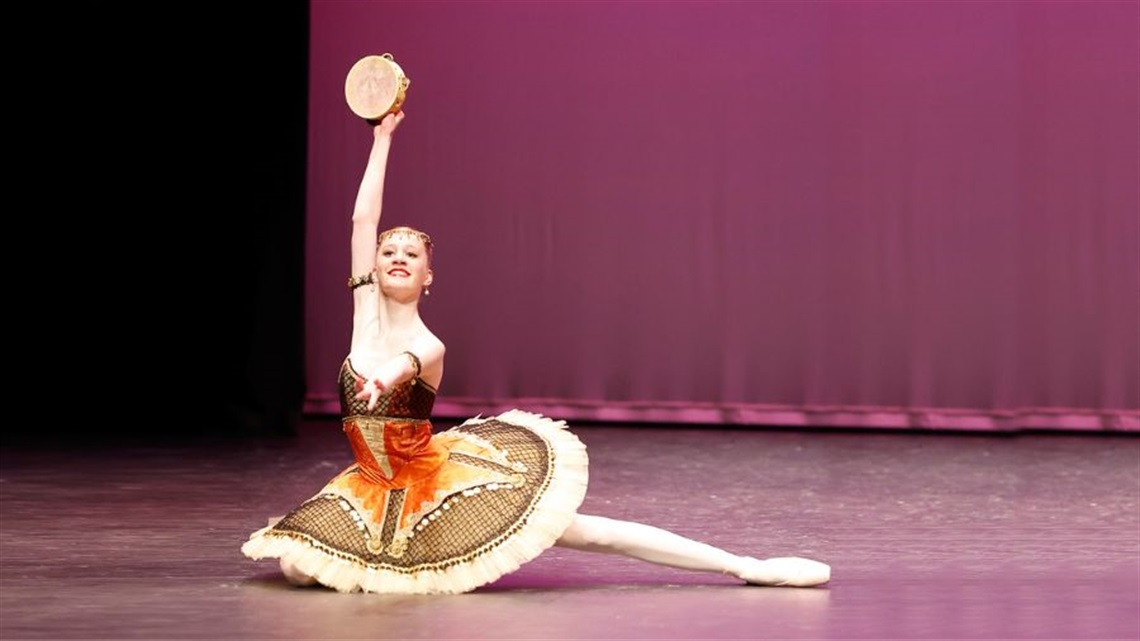 A ballerina dressed in a colourful tutu sits on a stage with one leg stretched out to the side. She raises a tambourine in air in her right hand.