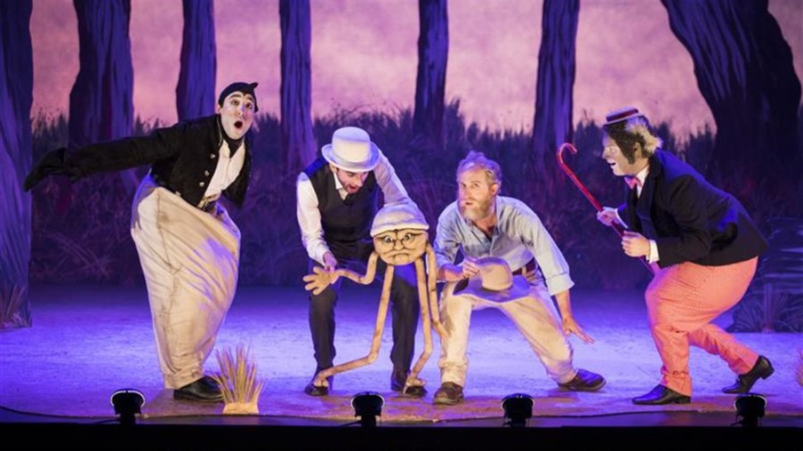 Four characters crouch next to each other across a stage. One holds a puppet with long legs. Another holds a red cane in their left hand.