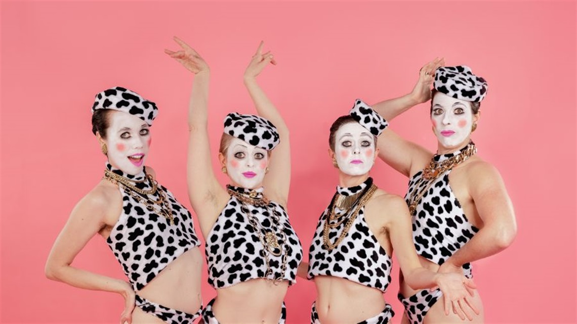 Four people wearing cow patch bikinis, matching hats and gold jewellery and clown makeup on their faces pose next to each other.