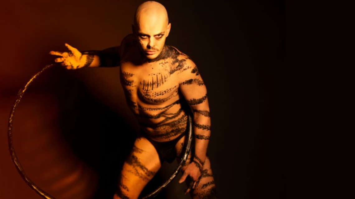 A bald-headed man holds a hula hoop in his criss-crossed hands around his upper body. He stands slightly crouching forward through the centre of the hula hoop. Black marks cover his nearly naked body. 