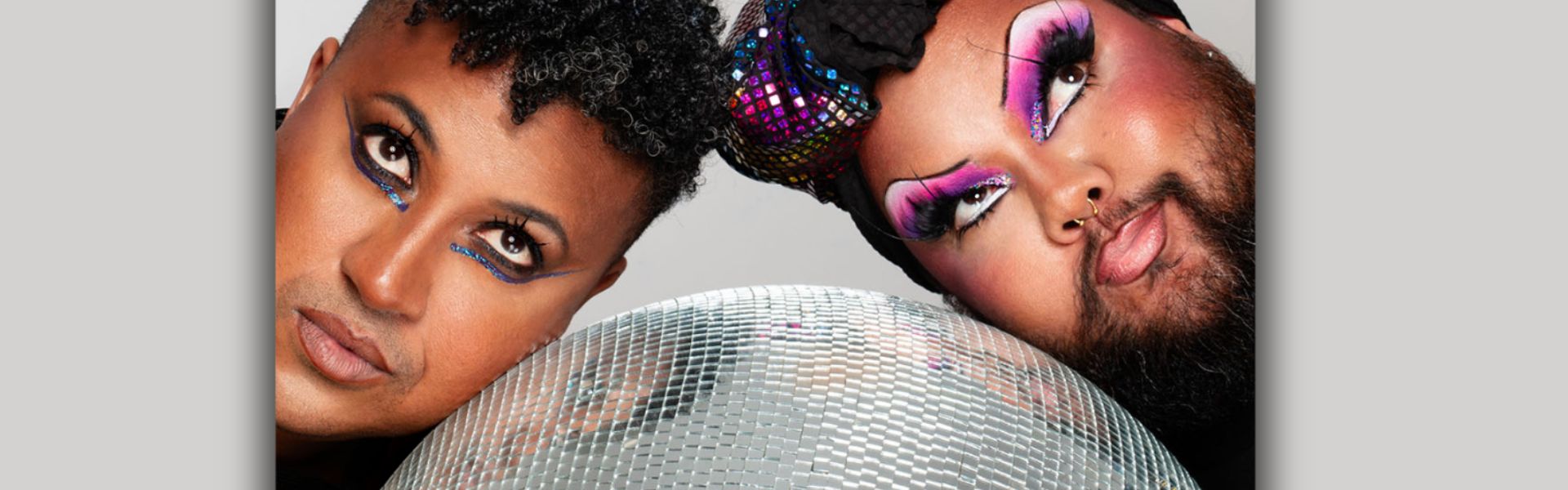 A close up of two people of colour wearing colourful makeup lay their heads gently on a mirror ball.