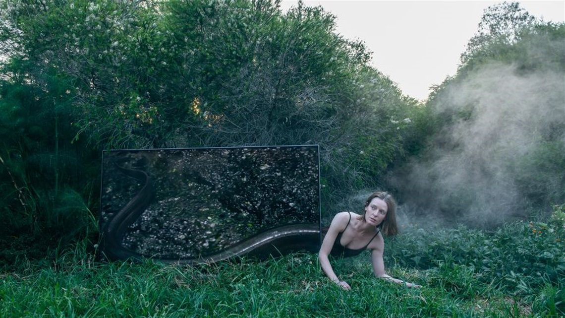 A woman perches herself on the grass in front of a digital screen so that her bottom half looks like an eel's tail.