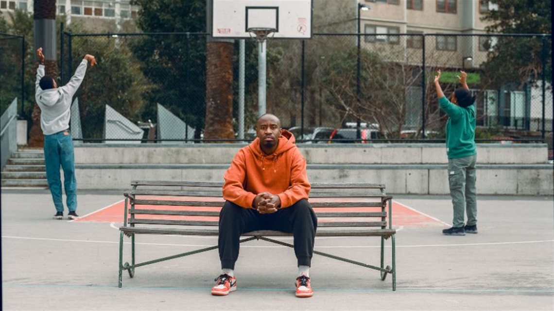 A Black man wearing a an orange jumper sits on a bench outdoors with his elbows resting on his thighs and hands folded. Behind him is an urban setting including a wire-fenced basketball court.