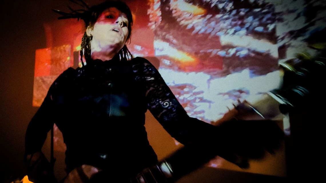 A person wearing dark clothing strumming a guitar in front of a projection.