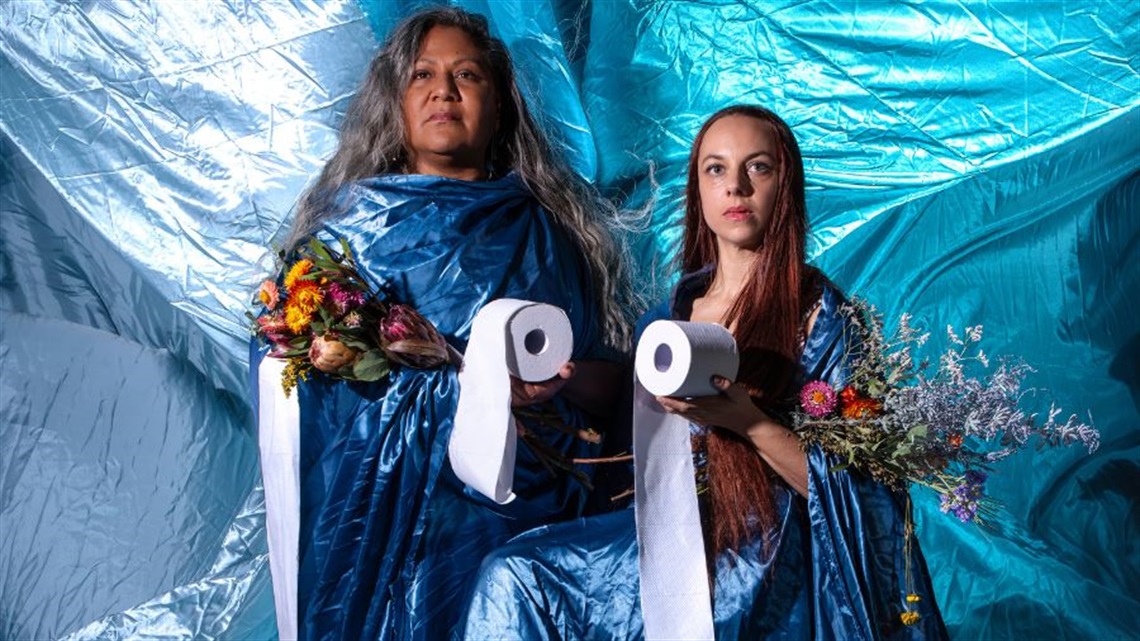 An Aboriginal woman with long dark hair stands behind a blue box, resting her hands on another woman with long dark hair lying on her back in the box with her head falling off the edge. They both wear Aboriginal garments. 