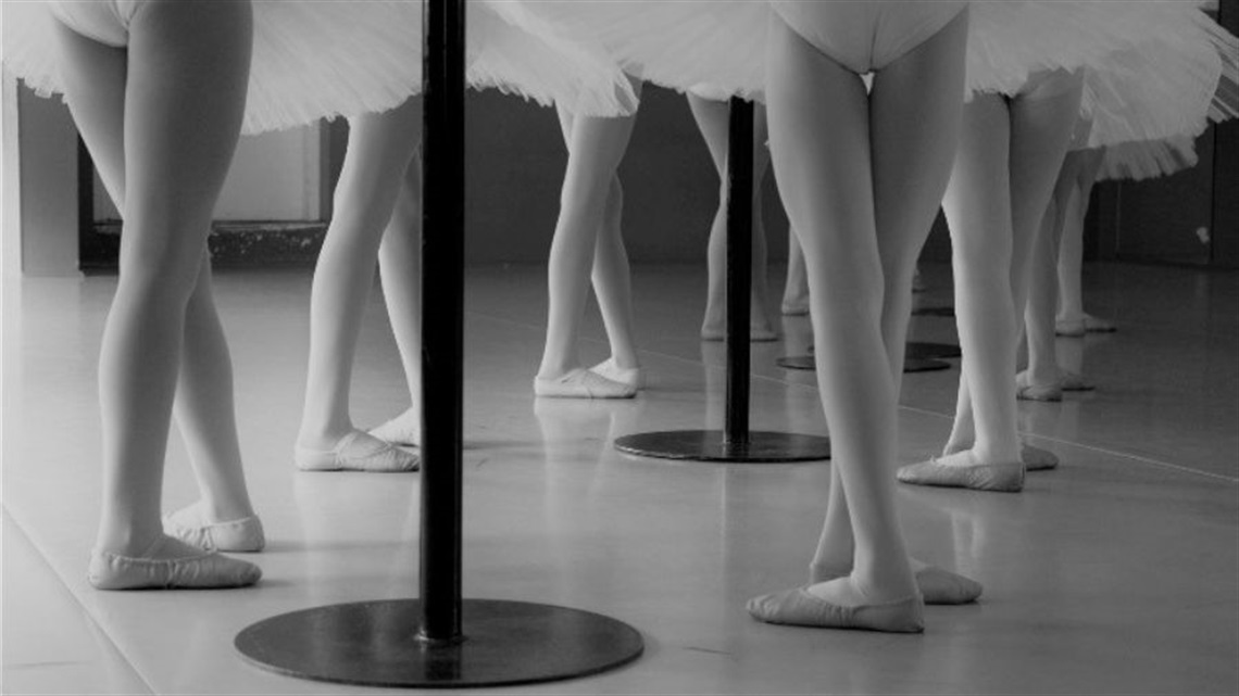 A black and white photograph of a cluster of ballet legs, wearing ballet slippers and tights. There is a black pole in the middle of the group.